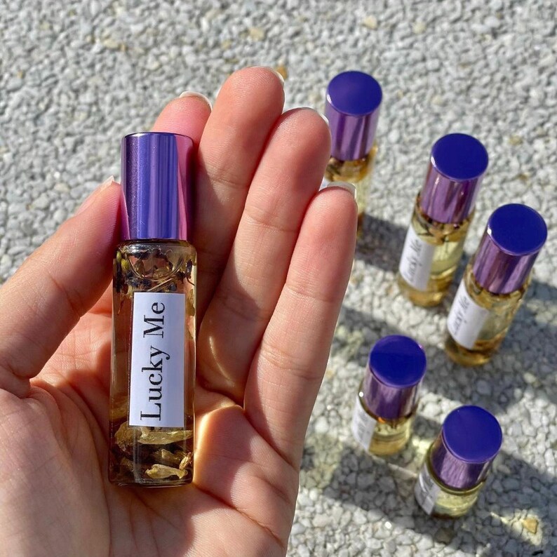 May include: A collection of small glass perfume bottles with purple caps. One bottle is held in a hand, displaying the "Lucky Me" label. The bottles contain a clear liquid with visible botanicals. The caps are a vibrant purple, and the bottles are arranged on a textured surface.