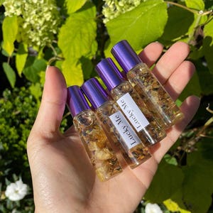 May include: A hand holds several small glass roll-on bottles filled with a clear liquid and dried botanicals. Each bottle has a purple cap and a label that reads "Lucky Me". The background features green foliage.