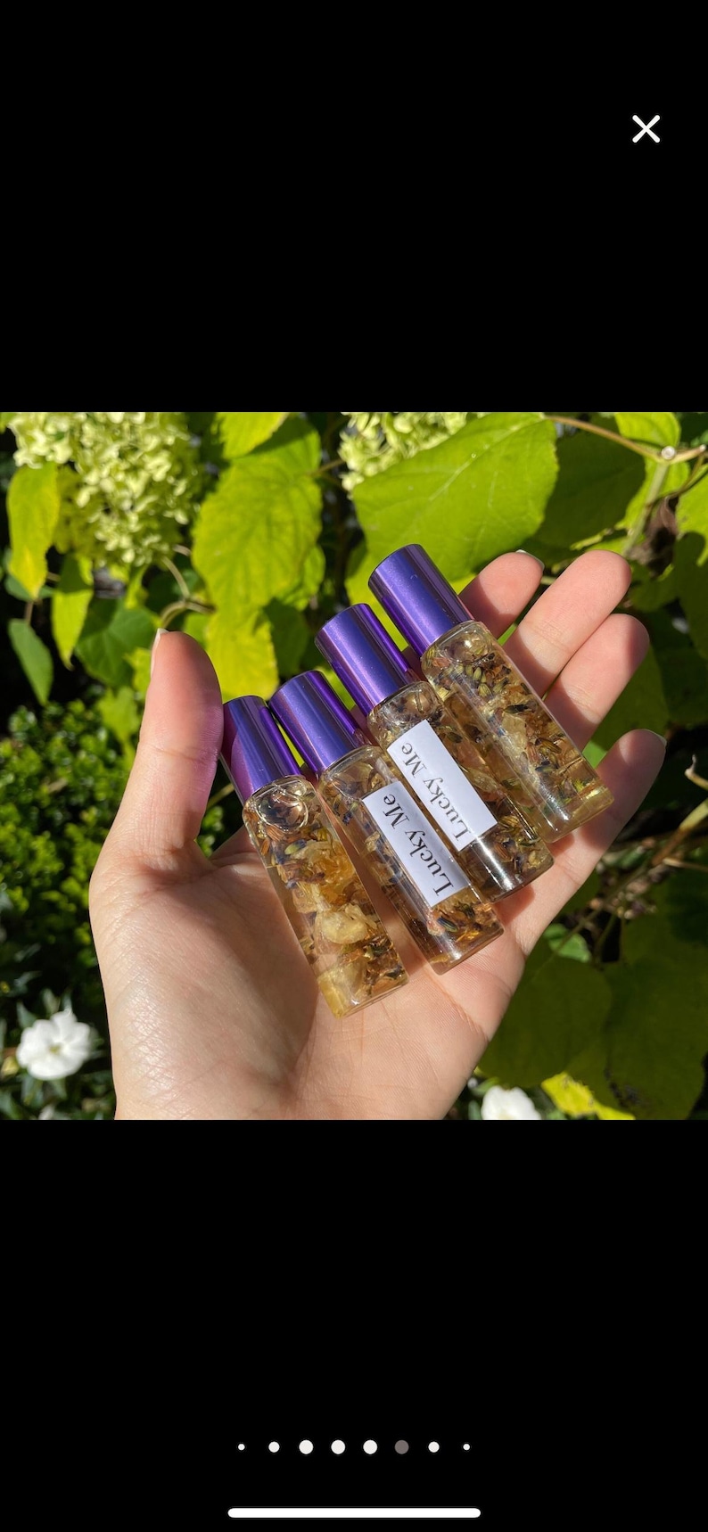May include: A hand holds several small glass roll-on bottles filled with a clear liquid and dried botanicals. Each bottle has a purple cap and a label that reads "Lucky Me". The background features green foliage.