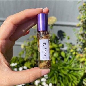 May include: A small glass essential oil roller bottle with a purple cap, filled with dried botanicals. The label reads "Lucky Me". The bottle is held in front of a blurred green background.