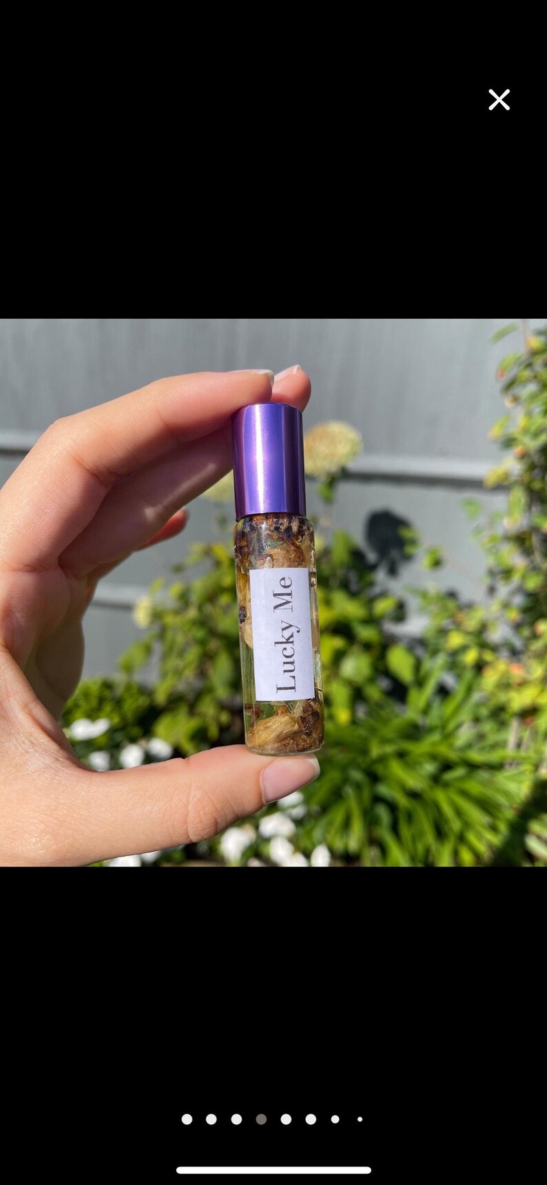 May include: A small glass essential oil roller bottle with a purple cap, filled with dried botanicals. The label reads "Lucky Me". The bottle is held in front of a blurred green background.