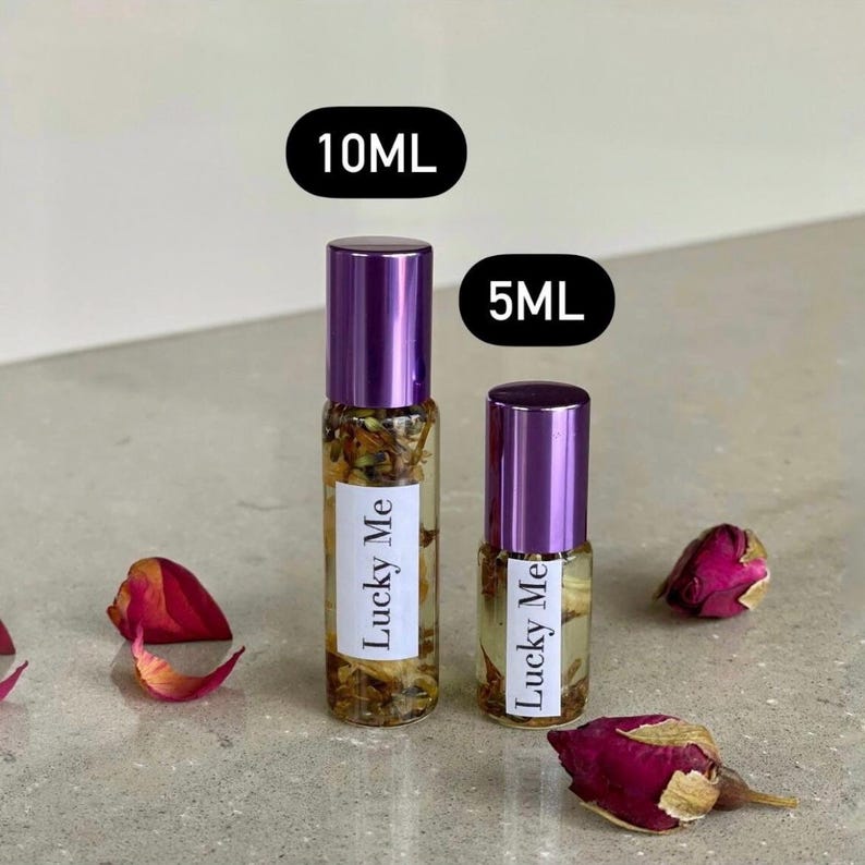 May include: Two glass roll-on bottles with purple caps, labeled "Lucky Me", are displayed. One bottle is labeled 10ML, the other 5ML. Dried flower petals and rosebuds are scattered around the bottles, creating a natural aesthetic.