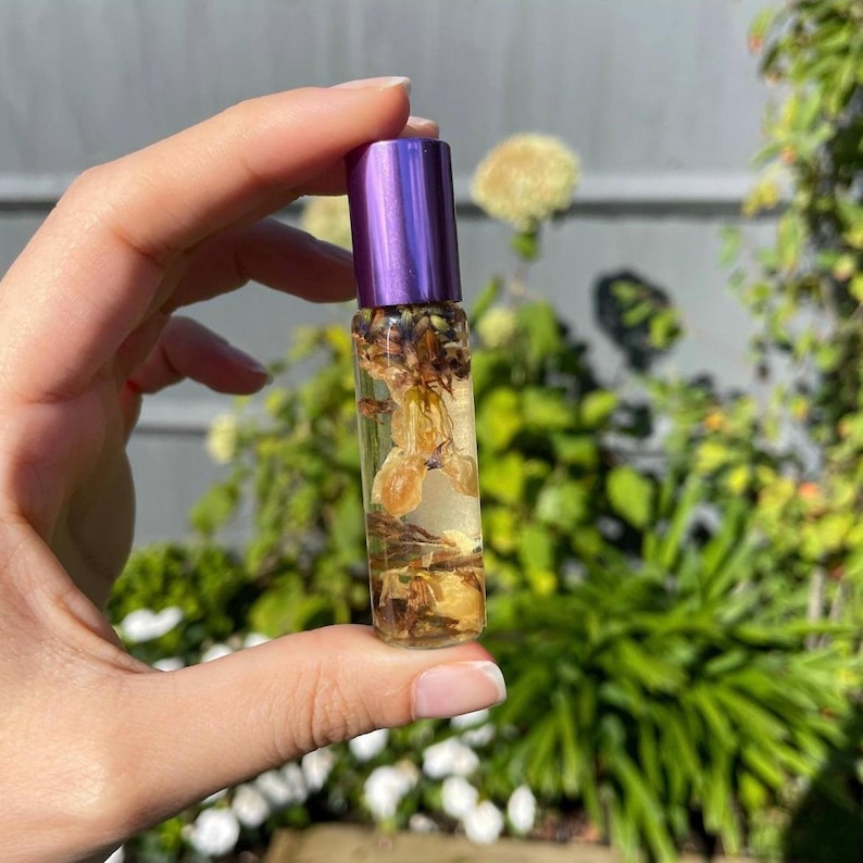 May include: A clear glass essential oil roller bottle with a purple cap. The bottle is filled with a light-colored liquid and dried flowers. The bottle is held in a hand against a blurred green background.