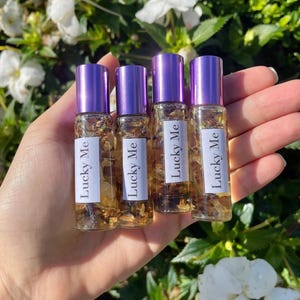 May include: Four small glass bottles with purple caps are held in a hand. Each bottle has a white label with the words "Lucky Me" printed on it. The bottles are filled with a golden liquid and small pieces of plant material.