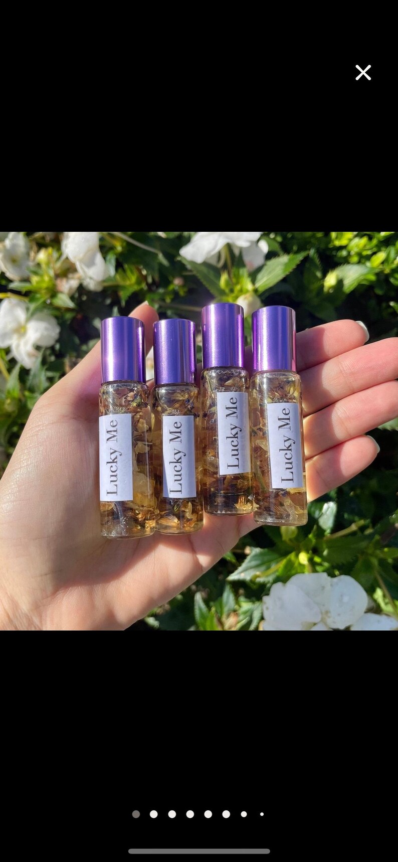 May include: Four small glass bottles with purple caps are held in a hand. Each bottle has a white label with the words "Lucky Me" printed on it. The bottles are filled with a golden liquid and small pieces of plant material.