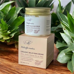 Könnte beinhalten: Ein Glas Midnight Mantra Organic Wild Yam Cream mit goldenem Deckel und weißem Etikett steht auf einer passenden Schachtel. Auf Schachtel und Glas befinden sich Text und ein Blumendesign. Das Produkt ist 60g.