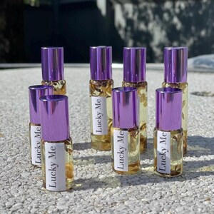 May include: A collection of small glass roll-on bottles with purple caps. Each bottle contains a clear liquid with dried botanicals and a label that reads "Lucky Me". The bottles are arranged on a textured surface.