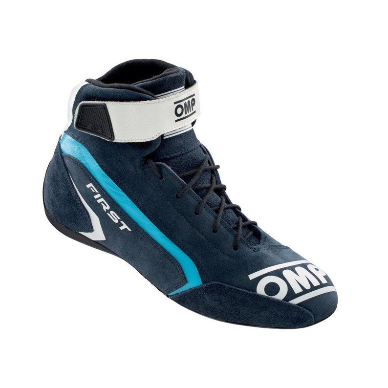 May include: Navy blue racing boot with a white strap and accents. The boot features the word "FIRST" in white and the brand name "OMP" in white. The boot has black laces and a light blue stripe.