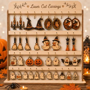 May include: A light-colored wooden display stand with the text Laser Cut Earrings, featuring four rows of hanging Halloween-themed earrings. Designs include black and orange witches' hats, owls, pumpkins, ghosts, ducks in hats, and brooms.