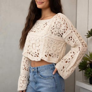 May include: Cream-colored crochet top with a square neckline and long bell sleeves. The top features a floral pattern and a cropped design. The model is wearing blue jeans. The top is made of a soft, open-weave material.