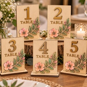 May include: A set of five rectangular wooden table number signs, numbered one through five. Each light wood sign features a large dark gold number, the word TABLE, and a decorative layered floral design with pink flowers and green leaves.