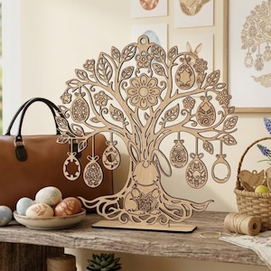 May include: A decorative wooden Easter tree with laser-cut designs, including a bunny and hanging egg decorations. The tree is displayed on a wooden surface, with a brown leather bag and a basket of eggs in the background. The tree is made of light-coloured wood.