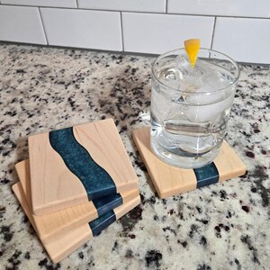 May include: Wooden square coasters with a teal river design. The coasters are made of light-colored wood with a dark teal epoxy resin inlay. A glass of water with ice and a lemon wedge sits on one coaster. The coasters are stacked on a speckled countertop.