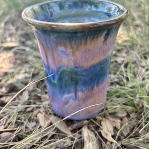 May include: A ceramic tumbler featuring a flared rim and a smooth, glossy glaze. The surface displays a blended color palette of deep blue, soft purple, and warm peach tones that transition vertically across the body of the vessel.