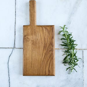 May include: A rectangular wooden chopping board with a handle, made of light brown wood. A sprig of green rosemary is placed to the right of the board. The board is on a white and grey tiled surface.