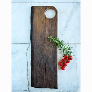May include: A dark brown wooden chopping board with a circular handle. A sprig of rosemary and a cluster of red cherry tomatoes are placed on the board. The board is on a white and grey tiled surface.