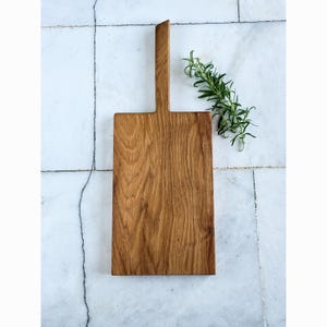 May include: A rectangular wooden chopping board with a handle. The board is made of light brown wood and has a natural wood grain pattern. A sprig of green rosemary is placed on the side of the board. The board is on a white and grey tiled surface.