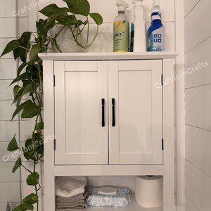 May include: A white bathroom cabinet with two doors and black handles. The cabinet has a shelf with towels, wipes, and toilet paper. Cleaning products sit on top, and a potted plant hangs on the side.