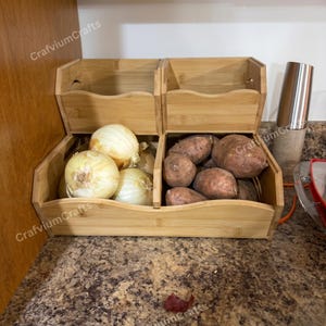 May include: A bamboo vegetable storage container with three compartments. The front compartment holds several yellow onions, and the middle compartment contains sweet potatoes. The container sits on a speckled countertop.
