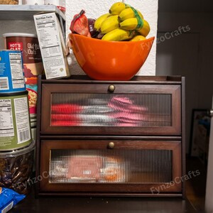 May include: A dark wood cabinet with two drawers, each with a glass front. The cabinet is topped with an orange bowl filled with bananas. Canned goods and boxes of oats are visible on the shelf above the cabinet.