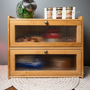 May include: A two-tiered bamboo bread box with glass-paneled doors. The doors have a ribbed texture and brass-colored knobs. The top of the box holds a glass jar and three small jars of preserves. The box sits on a woven placemat.