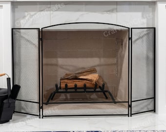 May include: A black metal fireplace screen with a mesh design, featuring a curved top and folding panels. Inside the fireplace, there are logs on a black metal grate. The fireplace is set against a white wall.