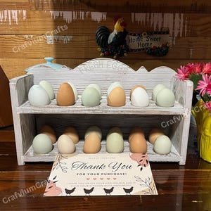 May include: A two-tiered, white-washed wooden egg holder filled with various colored eggs. The top shelf displays eggs in shades of white, brown, and green. The bottom shelf has similar eggs. A "Thank You" card is in front.