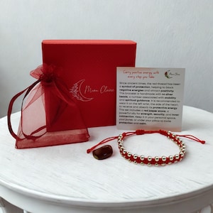 Puede incluir: Una pulsera roja con cuentas plateadas en un cordón rojo, acompañada de una piedra de jaspe rojo, una bolsa de organza roja, una caja de regalo roja con el logotipo de Moon Claire y una tarjeta informativa con texto sobre su significado.