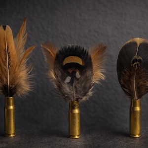 May include: Three decorative feather arrangements. Each feather is set in a gold-colored bullet casing. The feathers are brown, tan, and black. The background is a dark gray.