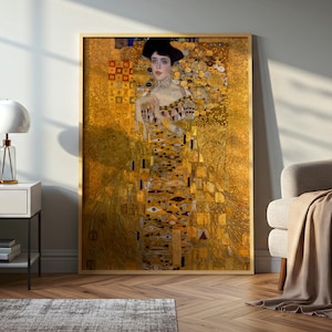 Op de afbeelding: Een ingelijste verticale kunstdruk van Gustav Klimts Portret van Adele Bloch-Bauer I. Het schilderij toont een persoon in een goudkleurige jurk versierd met complexe geometrische patronen, oogmotieven en metallic bladstructuren.
