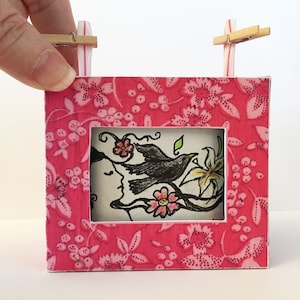 Op de afbeelding: Een klein roze frame met een bloemenpatroon, met een zwart-witte illustratie van een vogel en een bloem. Het frame wordt vastgehouden door twee houten wasknijpers die aan drinkrietjes zijn bevestigd.