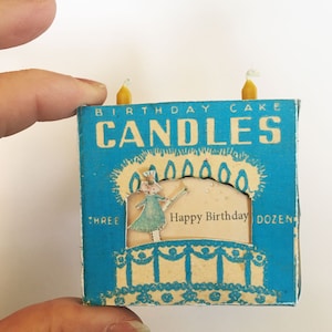 May include: A vintage blue box of birthday candles, with the words "BIRTHDAY CAKE CANDLES". The box has a cutout showing a cake with a fairy and the words "Happy Birthday". Two yellow candles are on top.