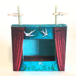 May include: A cardboard puppet theatre with a blue and green background, red curtains, and white birds. The theatre has a clear window for viewing puppets. Two straws are attached to the top of the theatre with clothes pegs.