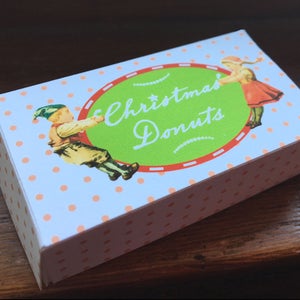 May include: A white box with pink polka dots and a green circle with the text "Christmas Donuts" in white. Two children are depicted on the box, one pulling a red ribbon attached to the circle.