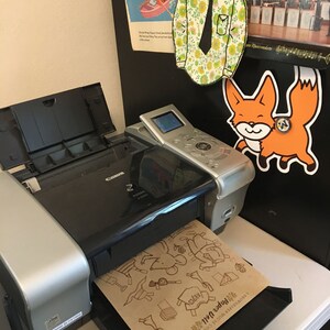 May include: A silver and black Canon PIXMA printer with a sheet of brown paper with a paper doll design in the paper tray. The paper doll design includes a fox, a house, a dress, a hat, and a pair of shoes. The text on the paper reads "A Brown Paper Doll Company, 2014,  Paper Doll"