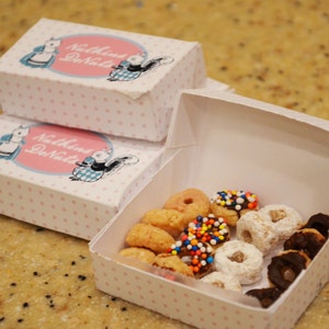 Könnte beinhalten: Eine weiße Box mit rosa Punkten und dem Text "Nuthin' But Donuts" darauf gedruckt. Die Box ist gefüllt mit einer Auswahl an Mini-Donuts, darunter einige mit Streuseln und Puderzucker.
