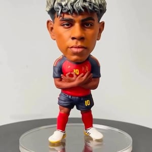 Puede incluir: Una figura cabezona de un jugador de fútbol con una cabeza grande y cabello texturizado de tono plateado. La figura viste una camiseta roja y azul oscuro con el número 10, pantalones cortos azul oscuro con el número 10, calcetines rojos y botas blancas.