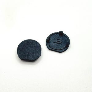 Op de afbeelding: Twee kleine, donkerblauwe, cirkelvormige plastic componenten. Eén is een effen schijf, en de andere heeft een textuur met een ontwerp en kleine lipjes. De items zijn tegen een witte achtergrond geplaatst.