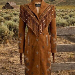 May include: A brown suede coat with fringe detailing on the shoulders and sleeves. The coat features intricate silver embellishments and a button-down front. The coat is displayed outdoors, with a rustic fence and a barn in the background.