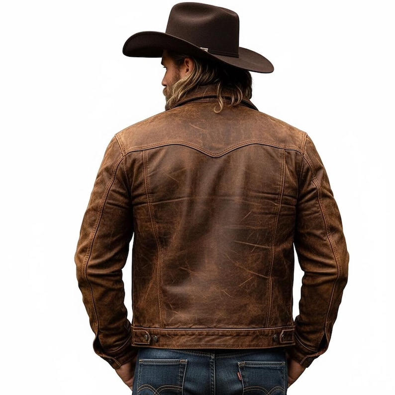 May include: A brown leather jacket with a distressed finish, featuring a classic collar and buttoned cuffs. The jacket has a western-style yoke detail on the back and is paired with blue jeans and a brown cowboy hat. The overall style is rugged and vintage.