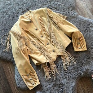 May include: A tan suede jacket with long fringe and decorative beading. The jacket features silver-toned buttons and is designed with a Western-inspired aesthetic. The fringe is along the sleeves and across the chest.
