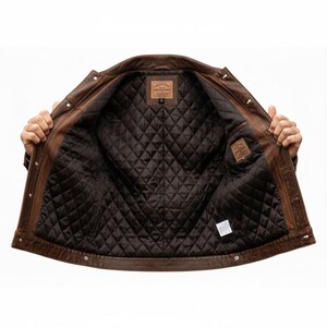 May include: Brown leather jacket with a quilted, dark brown interior. The jacket has a classic design with button closures and two small interior pockets. The jacket is open, revealing the interior lining.
