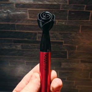Può includere: Una penna vape rossa e nera con una parte superiore a forma di rosa nera. La penna ha il testo "KIWI GO+" stampato sopra. Lo sfondo è un muro di mattoni scuri.