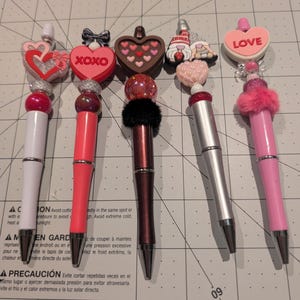 May include: A collection of decorative pens with various toppers. The pens feature designs such as hearts, bows, and the word "XOXO" and "LOVE". The pens have a variety of colors, including white, red, silver, and pink. The pens are arranged on a grid.