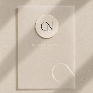Könnte beinhalten: Eine rechteckige beige Karte mit einem kreisförmigen, strukturierten Emblem, das die schwarzen Buchstaben CN trägt. Unter dem Emblem ist der Text CHIARA NAPOLI FASHION STUDIO in Weiß gedruckt. Ein großes, blasses weißes CN-Logo ist unten rechts.