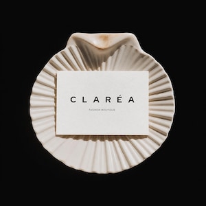 May include: A white seashell-shaped dish holds a business card with the text "CLARÉA FASHION BOUTIQUE" in black. The dish has a ridged design and sits against a black background, creating a minimalist aesthetic.