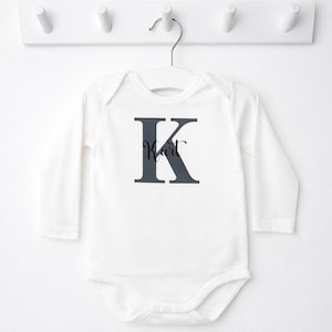 Könnte beinhalten: Weißer Baby-Body mit langen Ärmeln, mit einem großen marineblauen Buchstaben "K" und dem Namen "Kurt" in Schreibschrift. Der Body hängt an einem weißen Kleiderbügel.