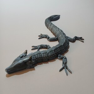 May include: A gray articulated dragon figurine with a textured, scaled body and four articulated legs. The dragon has a long, curved tail and a detailed head with horns. The figurine is made of a solid material.