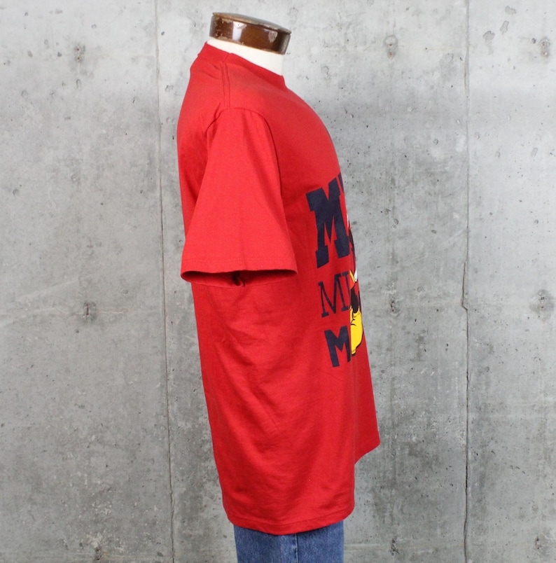 May include: A red t-shirt with the word "Mickey" in navy blue block letters and a yellow cartoon character. The shirt is made of a soft material and has short sleeves.
