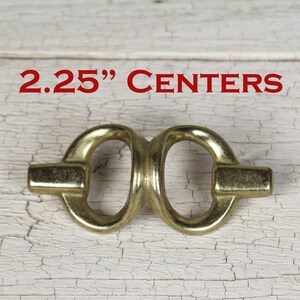 May include: A brass-toned metal drawer pull featuring two connected oval loops and horizontal end bars. The text 2.25" CENTERS is printed in red above the hardware, indicating the distance between the mounting points for this furniture handle.